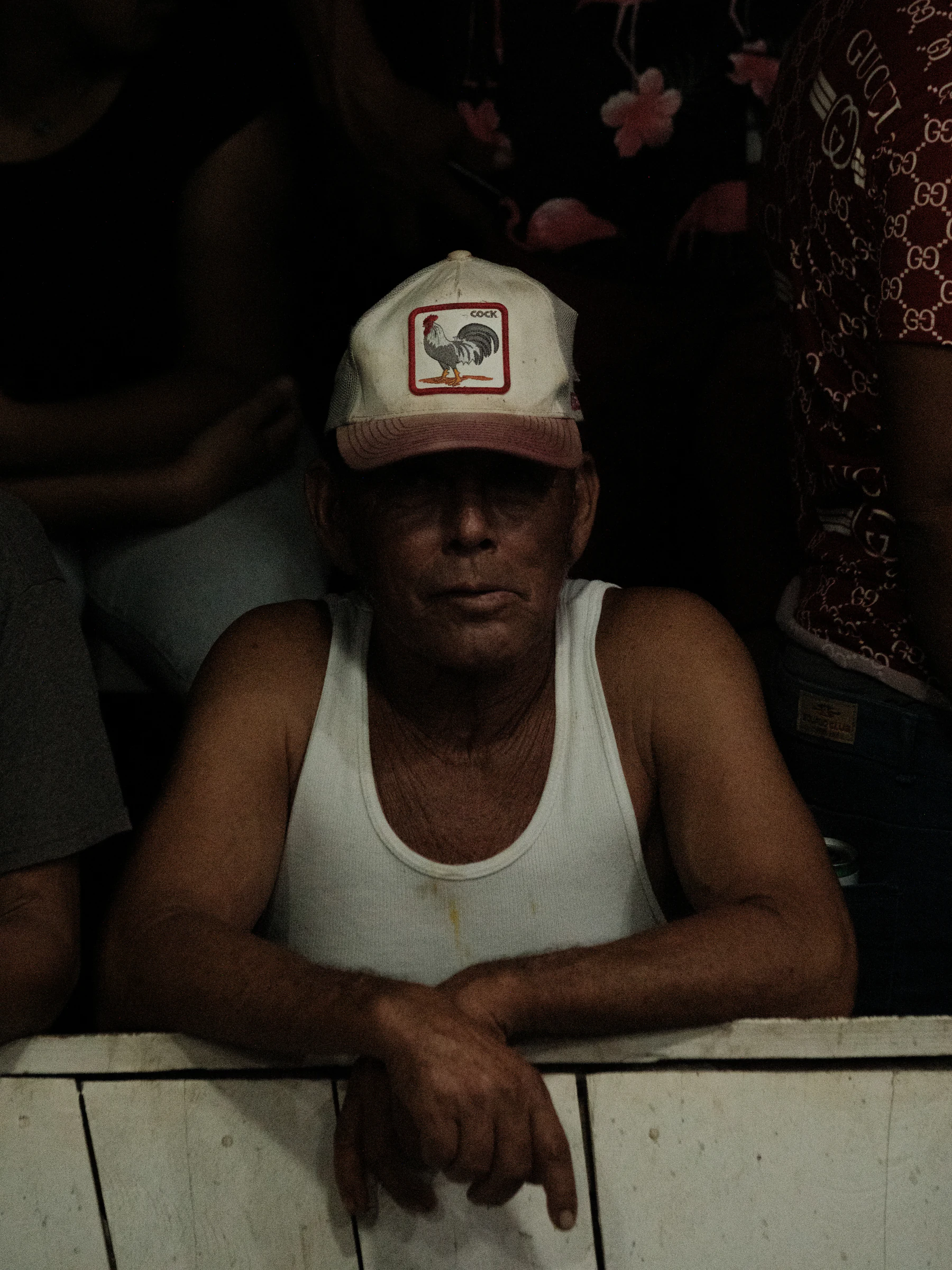 Man in a white tank top and rooster cap leaning over a barrier in low light