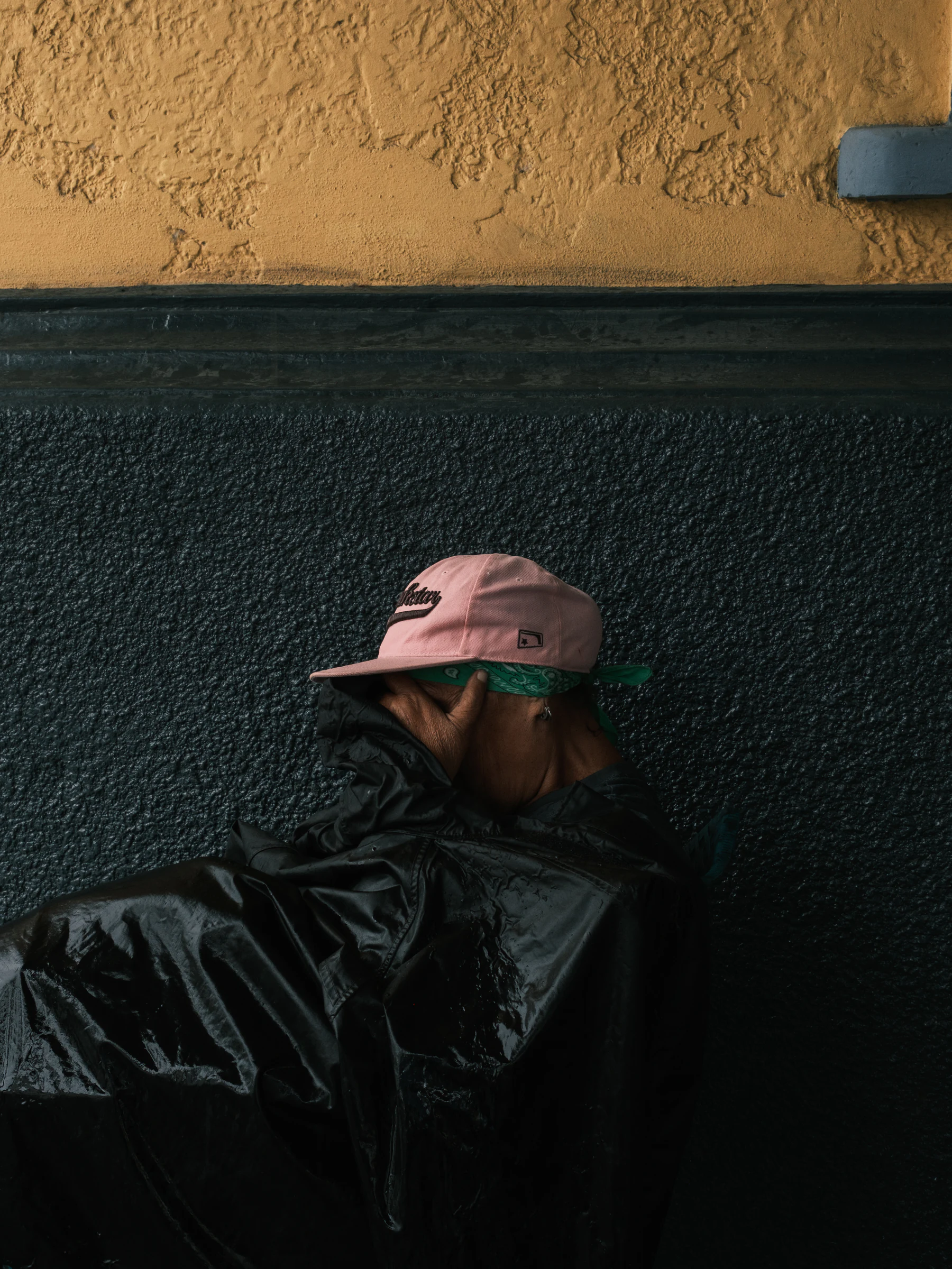 Person in a pink cap and dark rain jacket resting against a textured wall